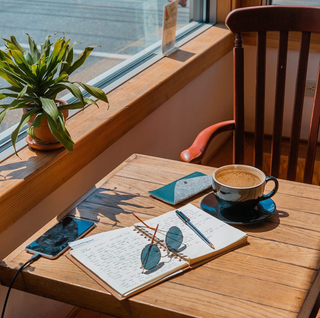 Coffee table at a cafe with a notebook, sunglasses, coffee and a phone that is charging.