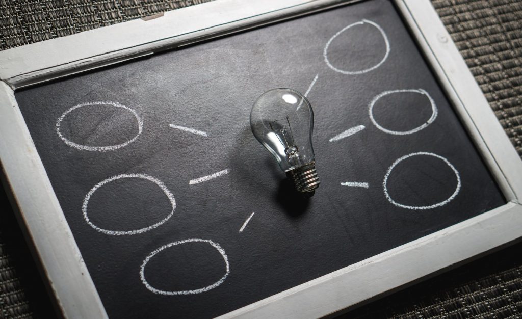 A lightbulb centered on a handheld chalkboard with empty circular thought bubbles branching off of it.
